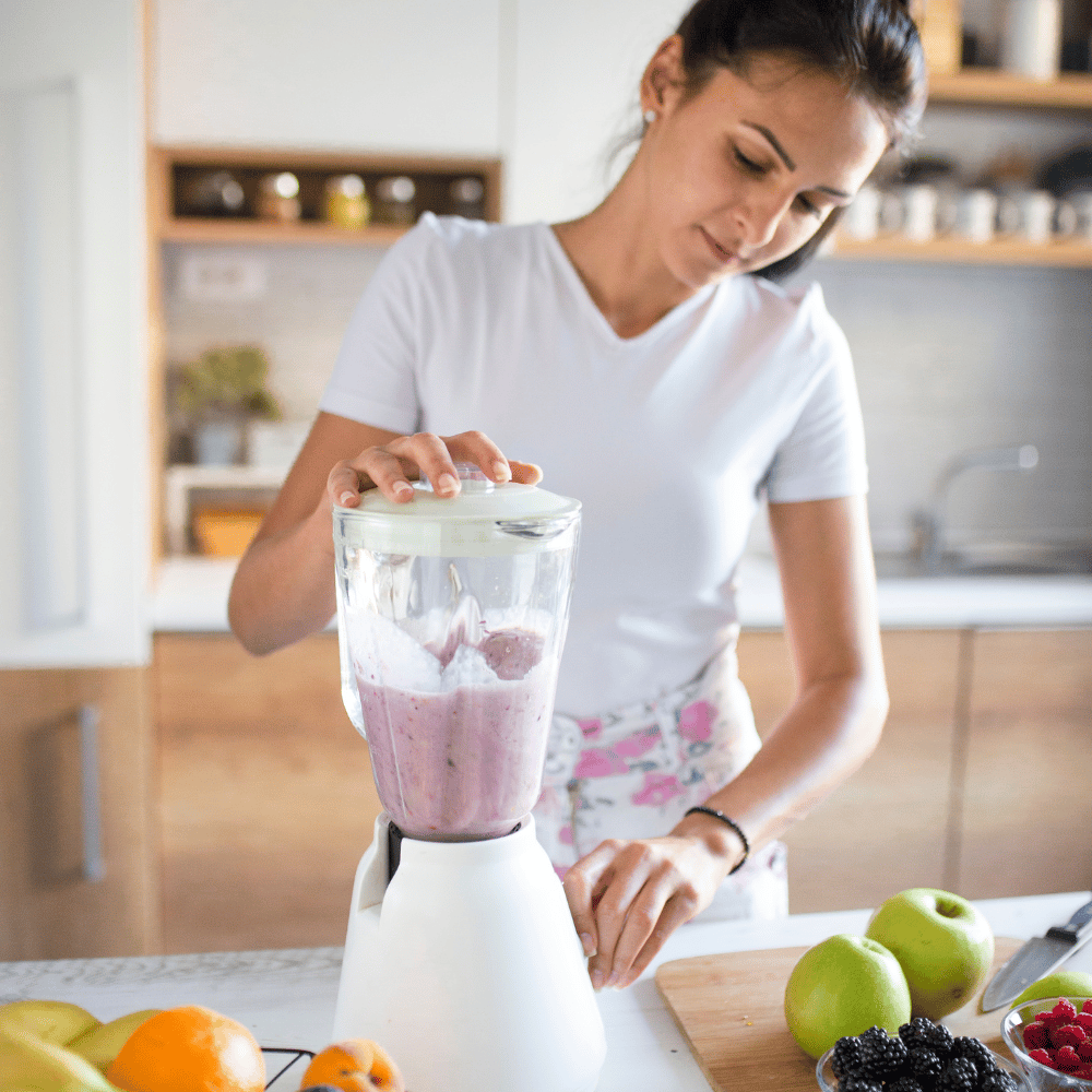 A woman in a white shirt with a dark head stands in the kitchen and blends a purple smoothie with her morning meal.