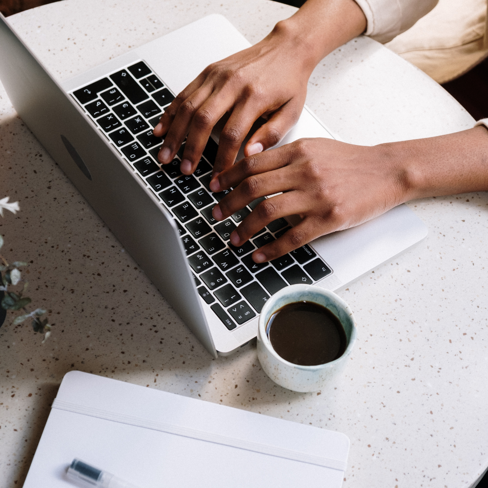 On the table is a laptop with a woman's hands writing on it, next to it is a cup of coffee.