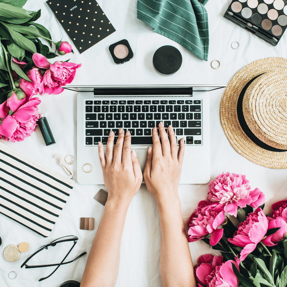 there is a laptop, roses, coffee on the white table and a woman's hands are typing on the laptop keyboard.