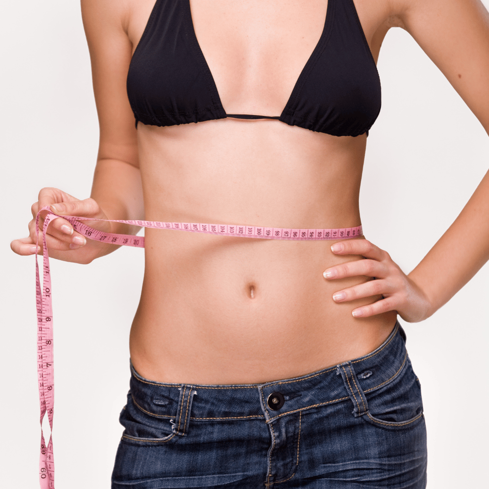 A woman stands on a gray background, wears a black bra, and measures her waist circumference with a tape measure.