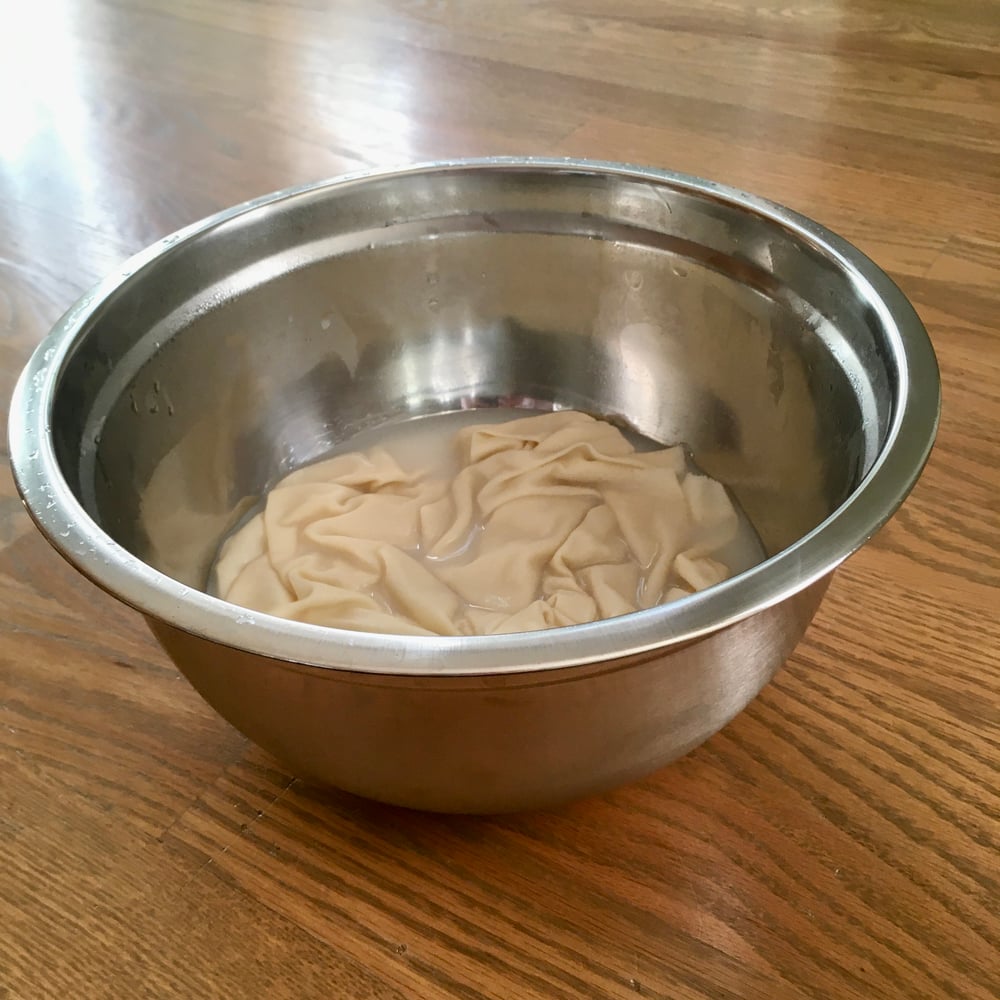 A stainless steel bowl containing light-colored fabric soaking in soy milk during the mordanting process for natural dyeing.