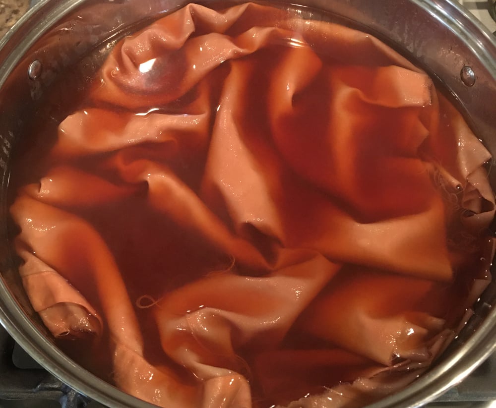 A stainless steel pot on a stove, filled with fabric immersed in a rich, maroon-colored natural dye made from avocado pits and skins. The fabric is evenly soaked, with folds visible above the liquid.