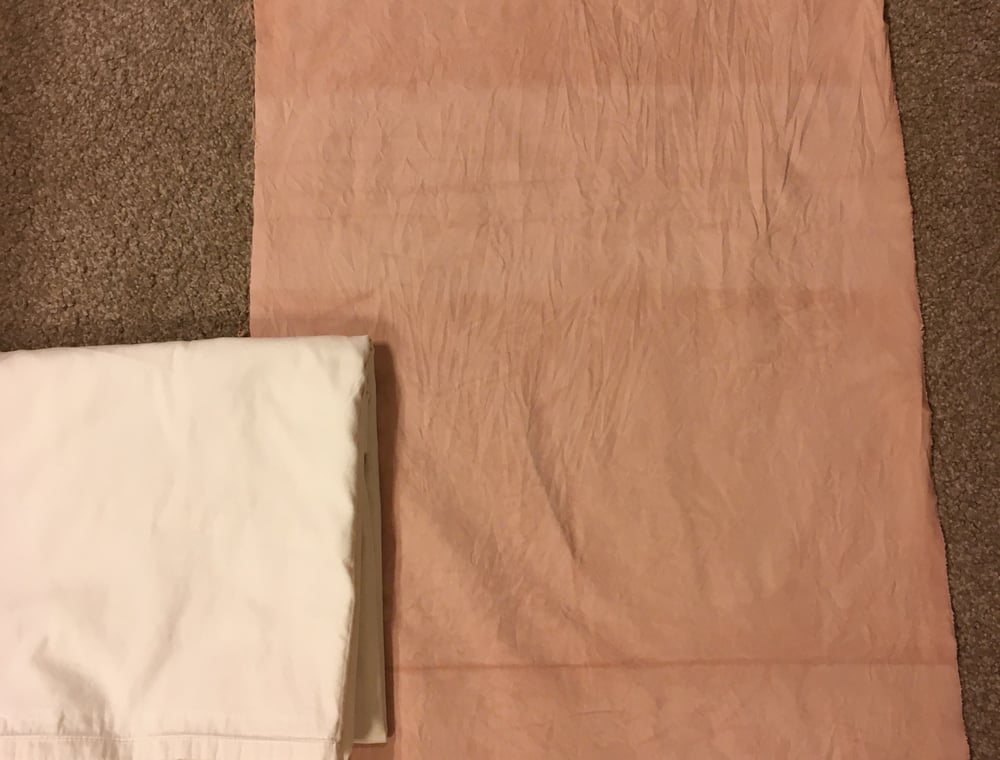 A side-by-side comparison of fabric before and after natural dyeing. On the left, the undyed fabric is white and neatly folded. On the right, the dyed fabric lies flat, showing a soft, pinkish-mauve hue achieved through the dyeing process. Both are placed on a beige carpet.