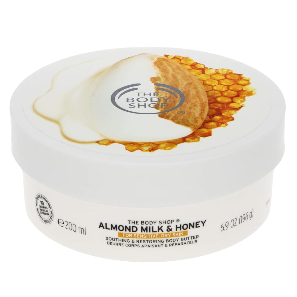 A white tub of The Body Shop Almond Milk &amp; Honey Body Butter, a nourishing body moisturizer for dry skin, featuring a lid with images of honeycomb and milk. The label indicates it is for sensitive, dry skin and the container holds 200 ml (6.9 oz).