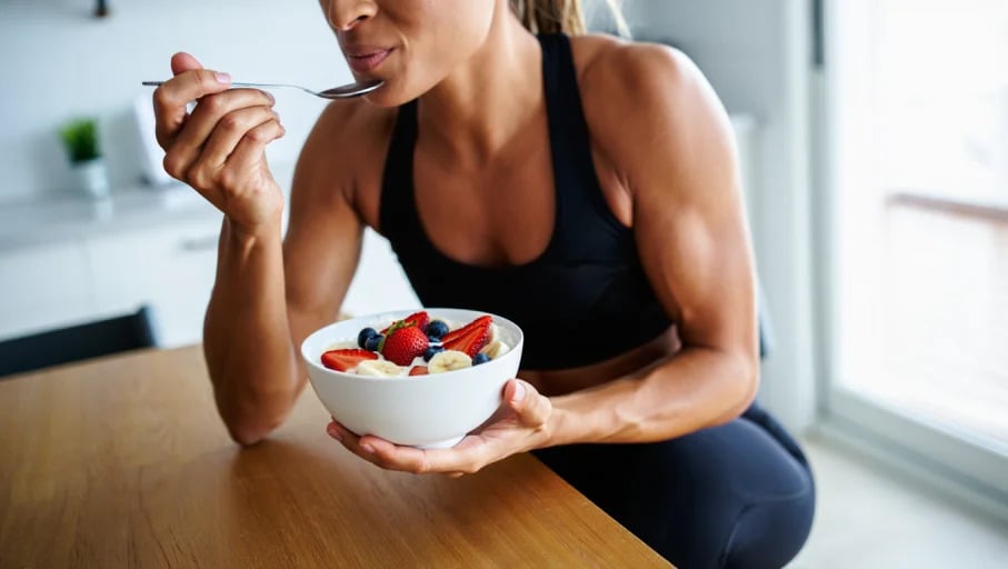 A fit woman in black athletic wear sits at a wooden table eating from a white ceramic bowl filled with Greek yogurt topped with fresh strawberries, blueberries, and sliced bananas. The woman holds a silver spoon in her right hand, bringing it towards her mouth. Her athletic build is evident through her form-fitting workout top and the definition visible in her arms. The scene takes place in a bright kitchen with white walls and natural lighting streaming through a window. Lifestyle photography with natural window lighting and shallow depth of field focusing on the subject in a bright indoor setting.