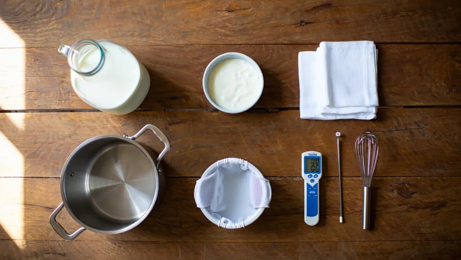 A flat lay of homemade Greek yogurt ingredients and tools: 1 gallon of organic whole milk in a glass jug, a small bowl of plain Greek yogurt starter, a large heavy-bottomed pot, an instant-read thermometer, a metal whisk, clean folded kitchen towels, and a yogurt strainer. Arranged neatly on a rustic wooden kitchen table, natural daylight, ultra realistic, high-resolution food photography, minimalist style --ar 16:9 --v 6 --q 2