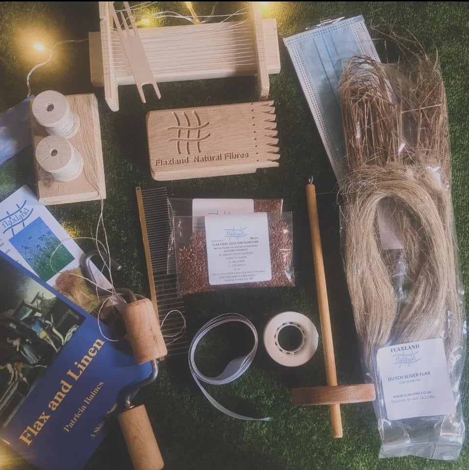 A collection of items associated with processing flax into linen. Top to bottom, left to right: a small wooden weaving loom, ready warped with linen thread, and shuttle. A paper mask. Two bobbins of linen thread on a bobbin holder. A wooden block with triangular teeth at one end. A packet of brown stalks. Some paper booklets, a metal-toothed comb. Two packets of brown seeds. A drop spindle. A packet of blonde fibres. A twine twisting device, a piece of ribbon, a roll of adhesive tape.