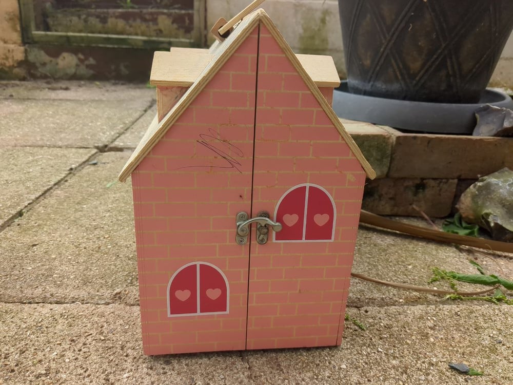 A side view of a doll's house bungalow with dormers on the roof. There is a latch holding the two halves of the house together, and false shuttered windows are printed on the wall.