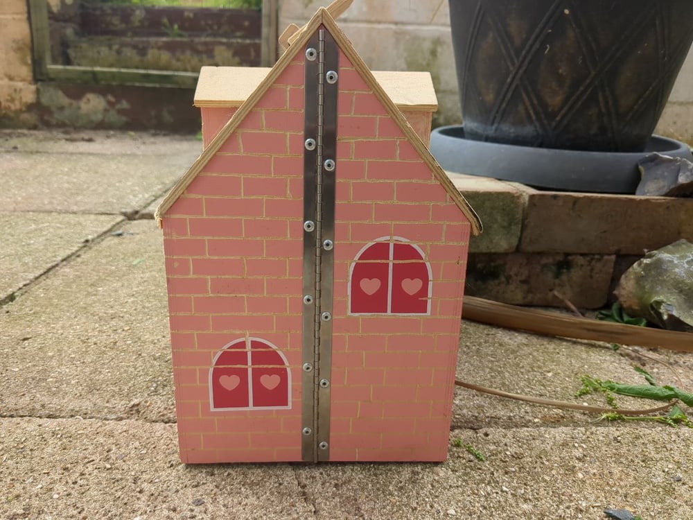 A bungalow type wooden doll's house with dormers; The brick details and two false shuttered windows are printed onto the wall in pink. There is a riveted hinge connecting the two halves of the house.