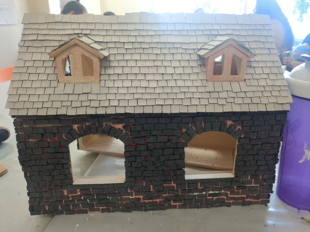 A bungalow doll's house with roof dormers, the wall is covered in scruffy black bricks and the roof is tiled with grey cardboard pieces like roofing slates.
