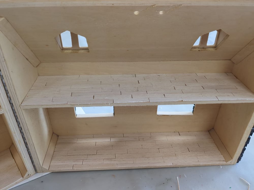 An inside view of a wooden doll's house. Both the bottom floor and the attic with dormers have been floored in strips of wood