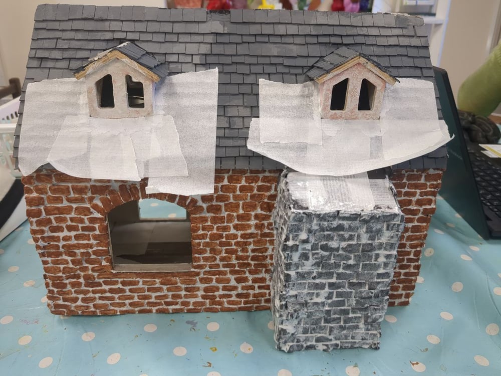 An outside view of a single storey doll's house with attic dormers. An unpainted but brick-textured chimney breast has been built out from one of the ground floor windows, ending just below the roof tiles in line with the dormer window. Masking tape surrounds the dormer windows, which have been painted with a white textured paint.
