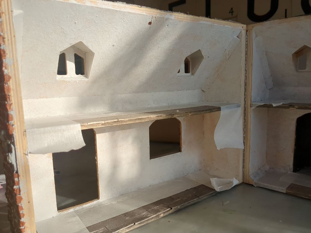 An inside view of a doll's house. The floor has been partially masked off and the walls and ceilings have been painted in a textured white paint.