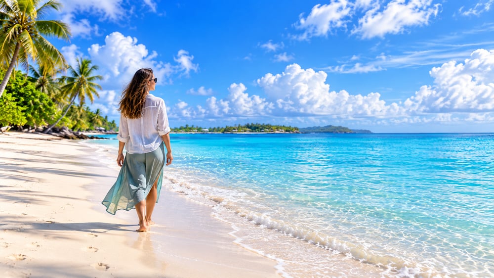 Woman walking thoughtfully along a quiet tropical shoreline beside clear turquoise water under an expansive blue sky.
