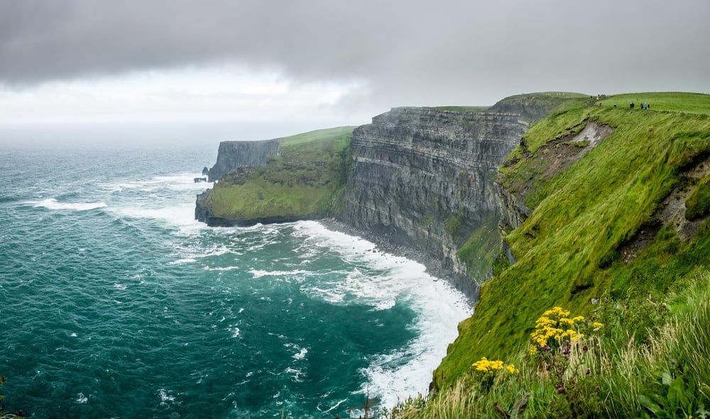 stock photograph of Irish coast showing grey rock cliffs and lusciously green fields with some yellow blooms in front, sky is overcast with grey clouds and the ocean water is blue with white foam lapping at the land