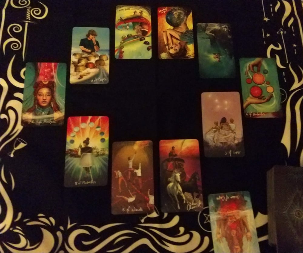 photograph taken by Evylyn Rose of a darkly-lit Tarot spread; cards are on an elemental-themed black and white cloth, laid out in an eye-shaped oval with an extra card off to the bottom right; Deck used is The Lightseer's Tarot by Chris-Anne
