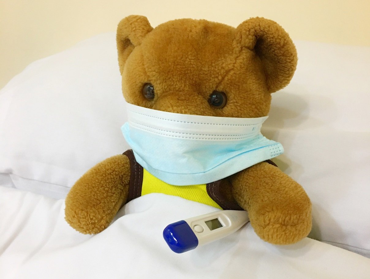 Teddy bear wearing a surgical mask, in bed with a thermometer under its arm. Photo by Kristine Wook on Unsplash