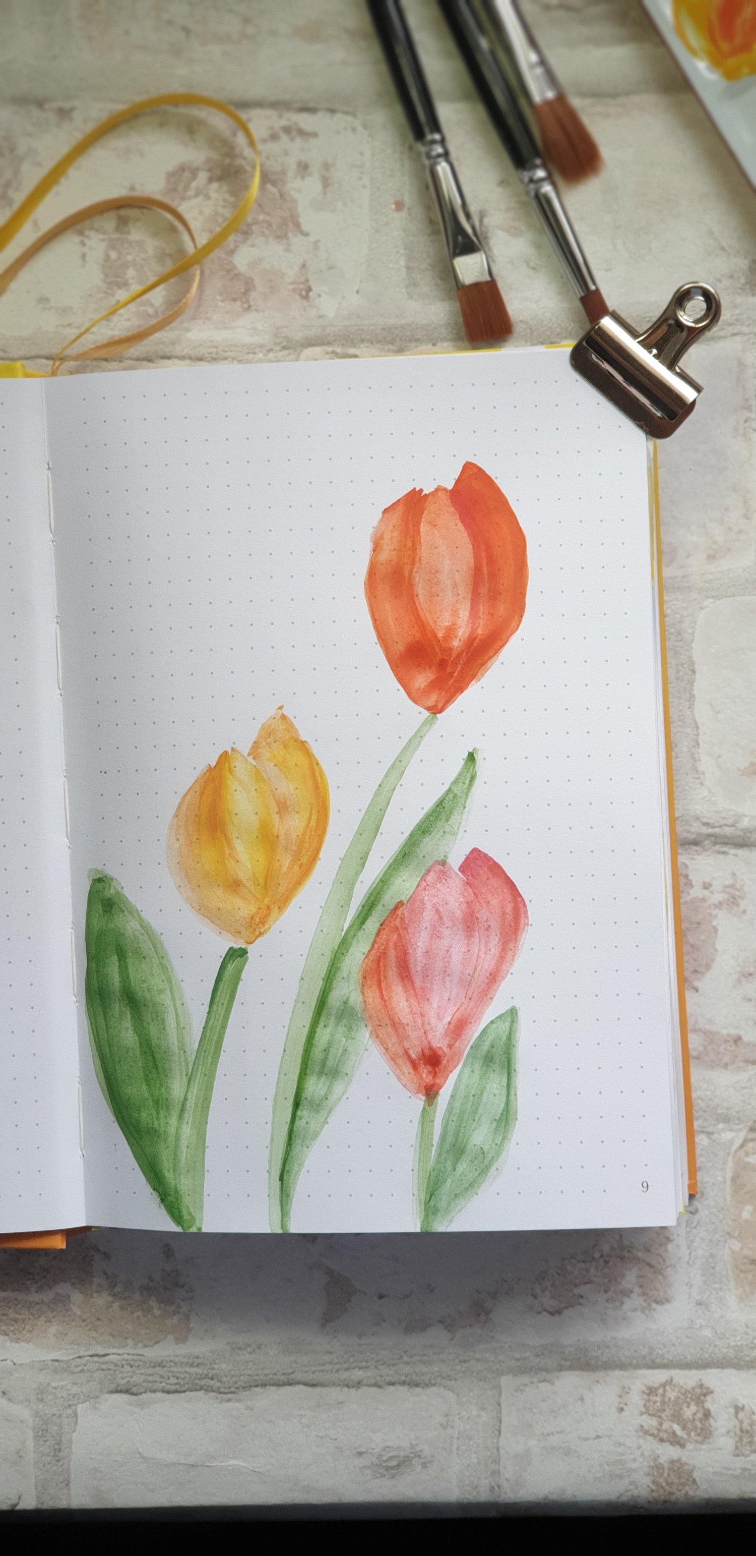Painting in a journal of three tulips in yellow, red and pink, on a brick effect background with three paintbrushes.