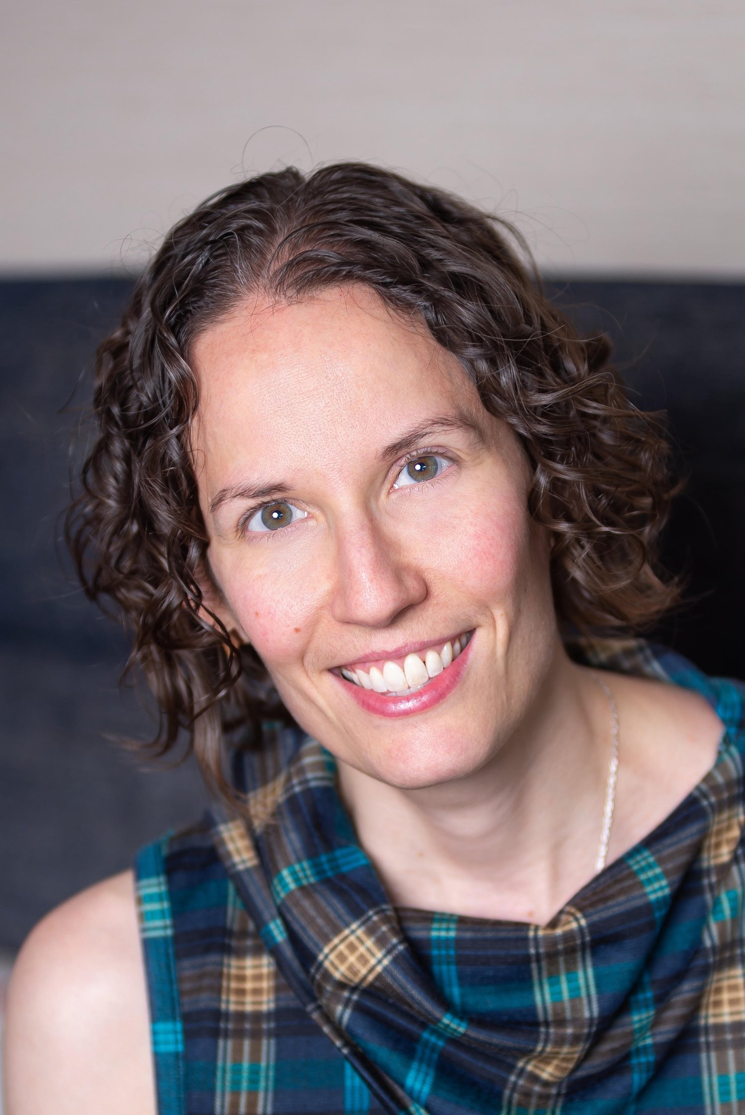 Headshot of Rose Prendeville. She's wearing a blue and teal and gold plaid dress with a scarf-like neckline and she has short curly hair.