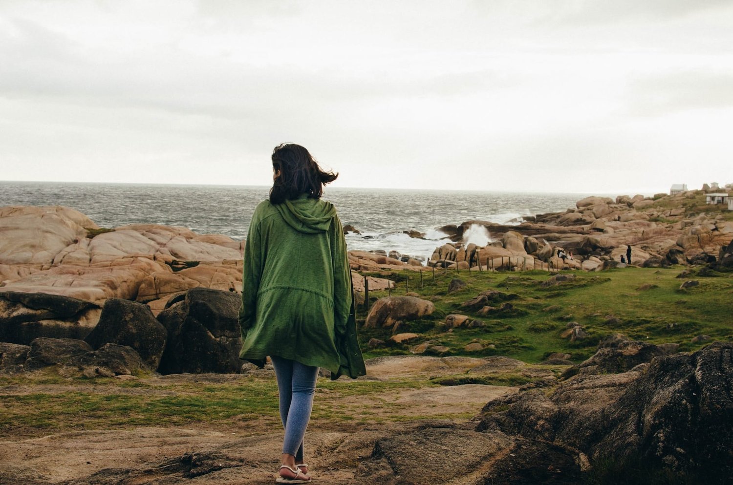 women walking alongside the ocean cliffs in jeans and a long green jacket for article on walking benefits.