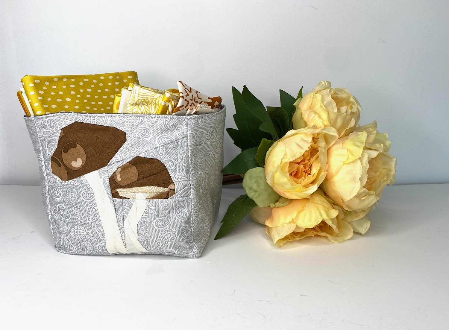 This cute Storage box is made using fabric scraps and an orphan quilt block with the picture of a mushroom on it. The storage box has been filled with pretty quilting fabrics and is sitting next to a bunch of paper roses.