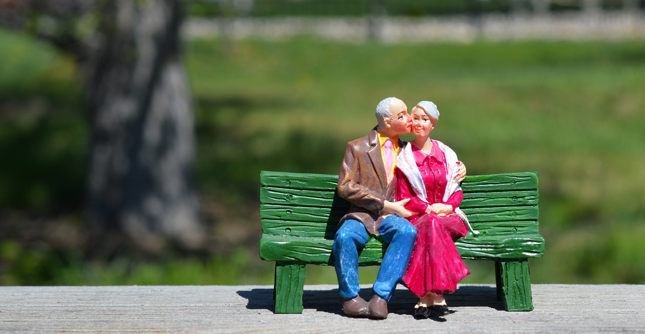 Foto de figurita de porcelana de pareja de ancianos sentados en un banco verde abrazados y dándose un beso en medio del parque. El señor tiene pelo corto y gris, y viste una americana marrón oscuro, camisa naranja, y unos vaqueros. La señora tiene pelo gr