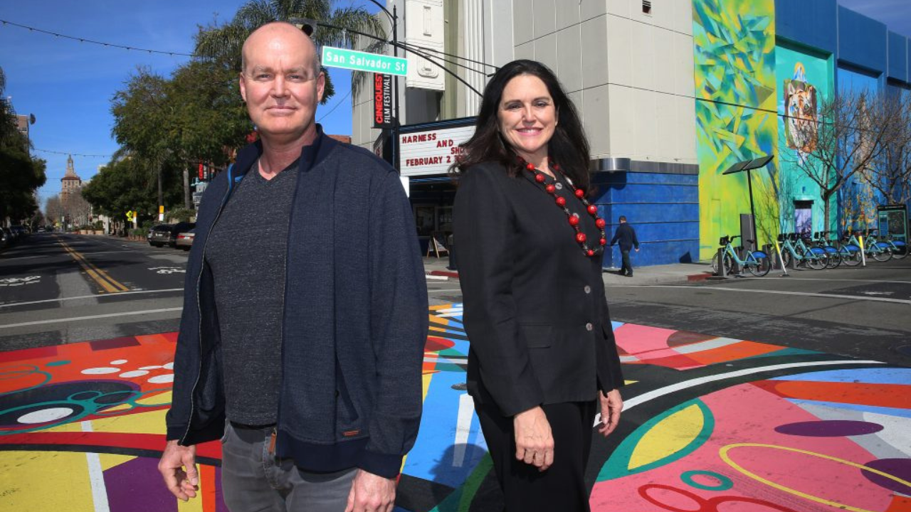 A white bald man and a white brunette woman standing next to each other against a very colorful pavement background