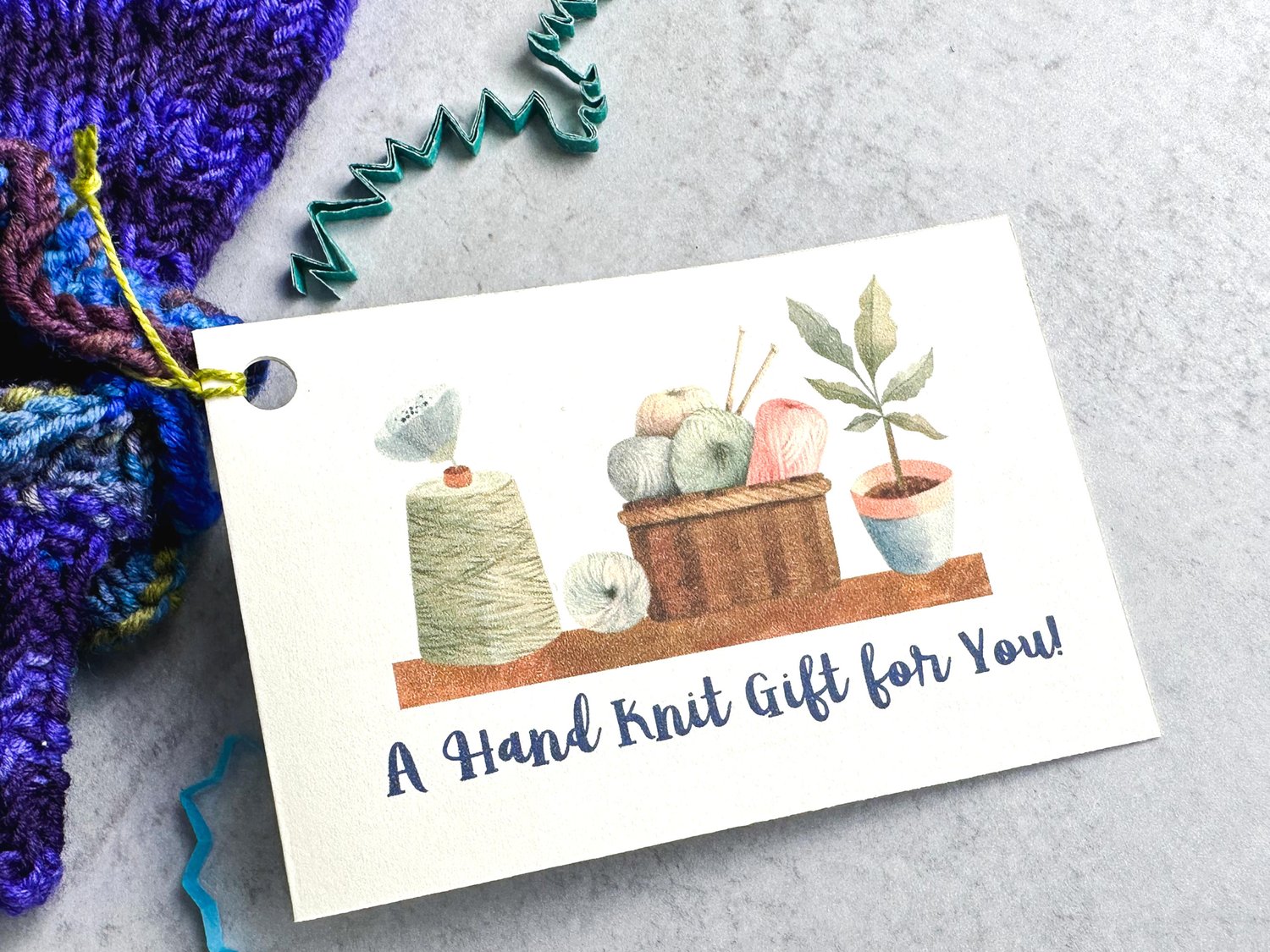 Card with watercolor painting of knitting yarns and a plant on a shelf attached to dark purple knitted item on a light grey background