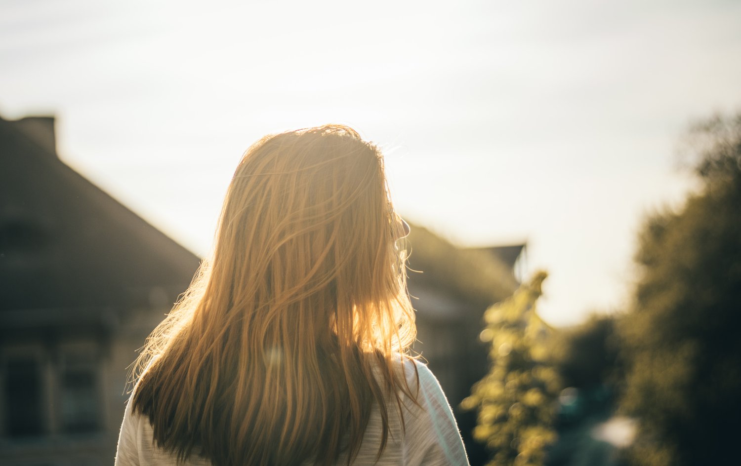 A blond Caucasian woman stand with her back to the camera looking over a neighbourhood late in a sunny day.