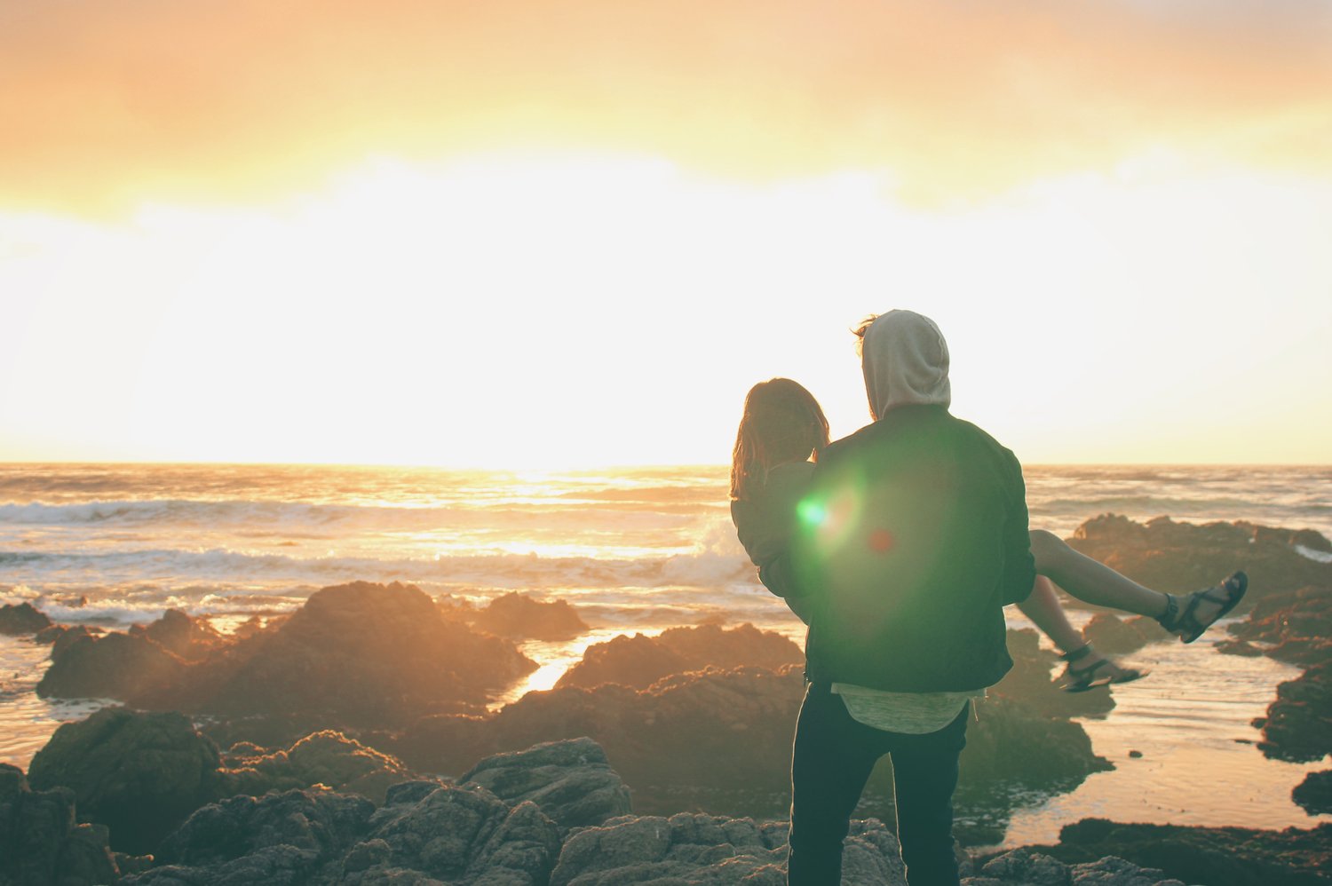 A man carries his girlfriend in his arms as they both look over the ocean at sunset.