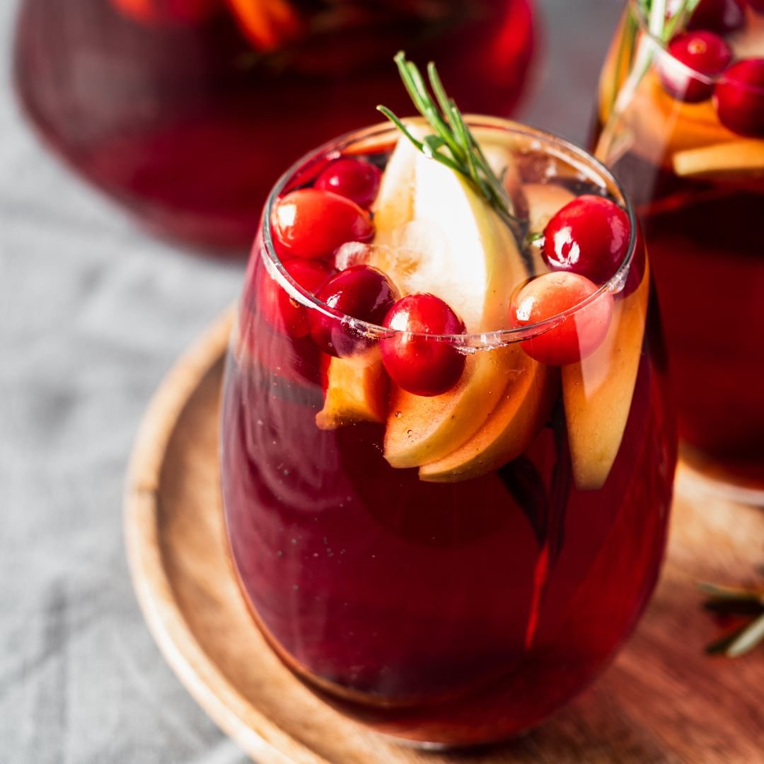 A festive glass of winter sangria featuring rich red wine, vibrant seasonal fruits, and aromatic spices. The drink is garnished with rosemary, apple slices and fresh cranberries, creating a visually appealing and flavorful holiday beverage.