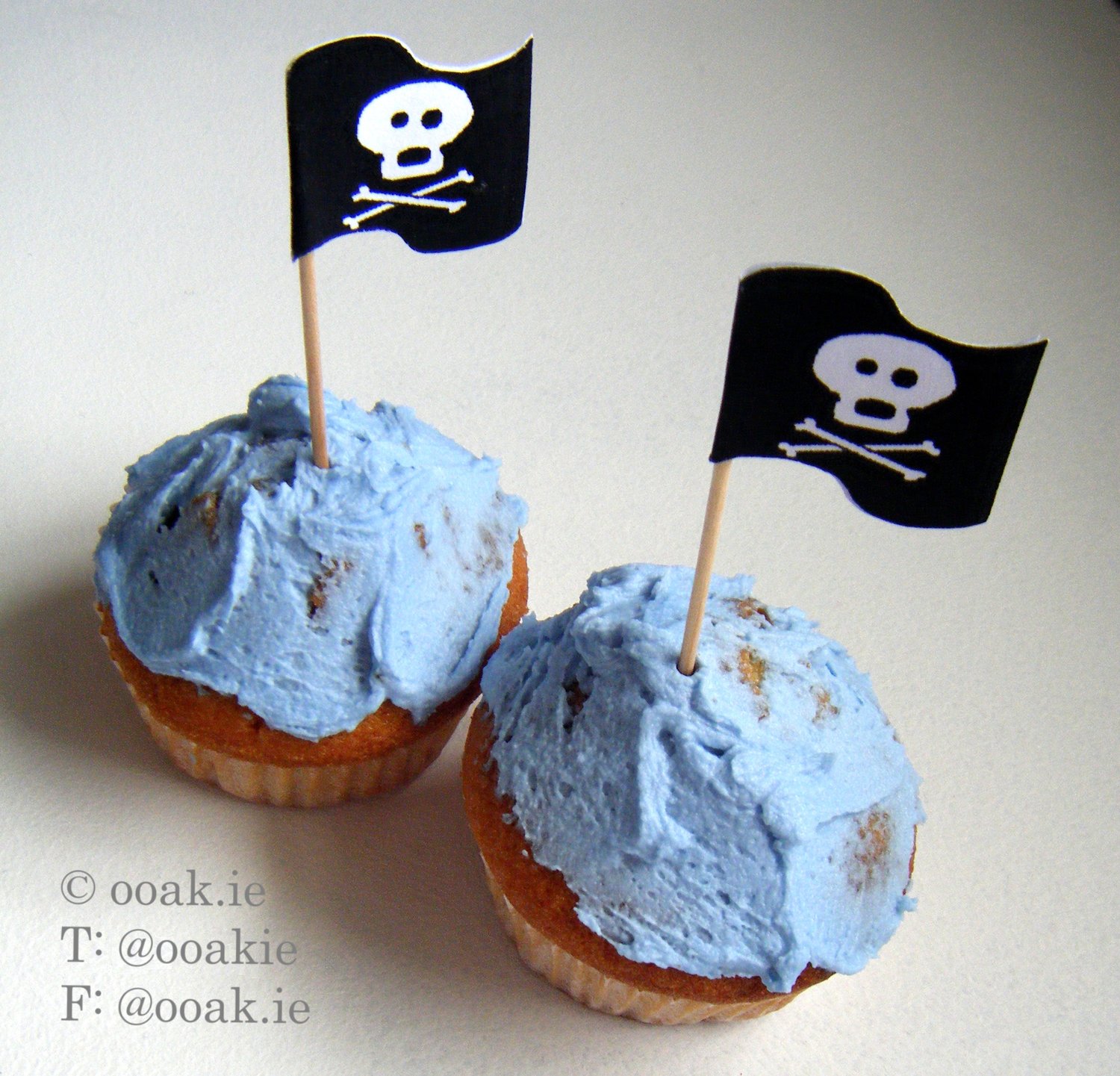 Printable sheet of 25 black and white Jolly Roger pirate flags with skull and crossbones, shown as DIY cupcake toppers on toothpicks.