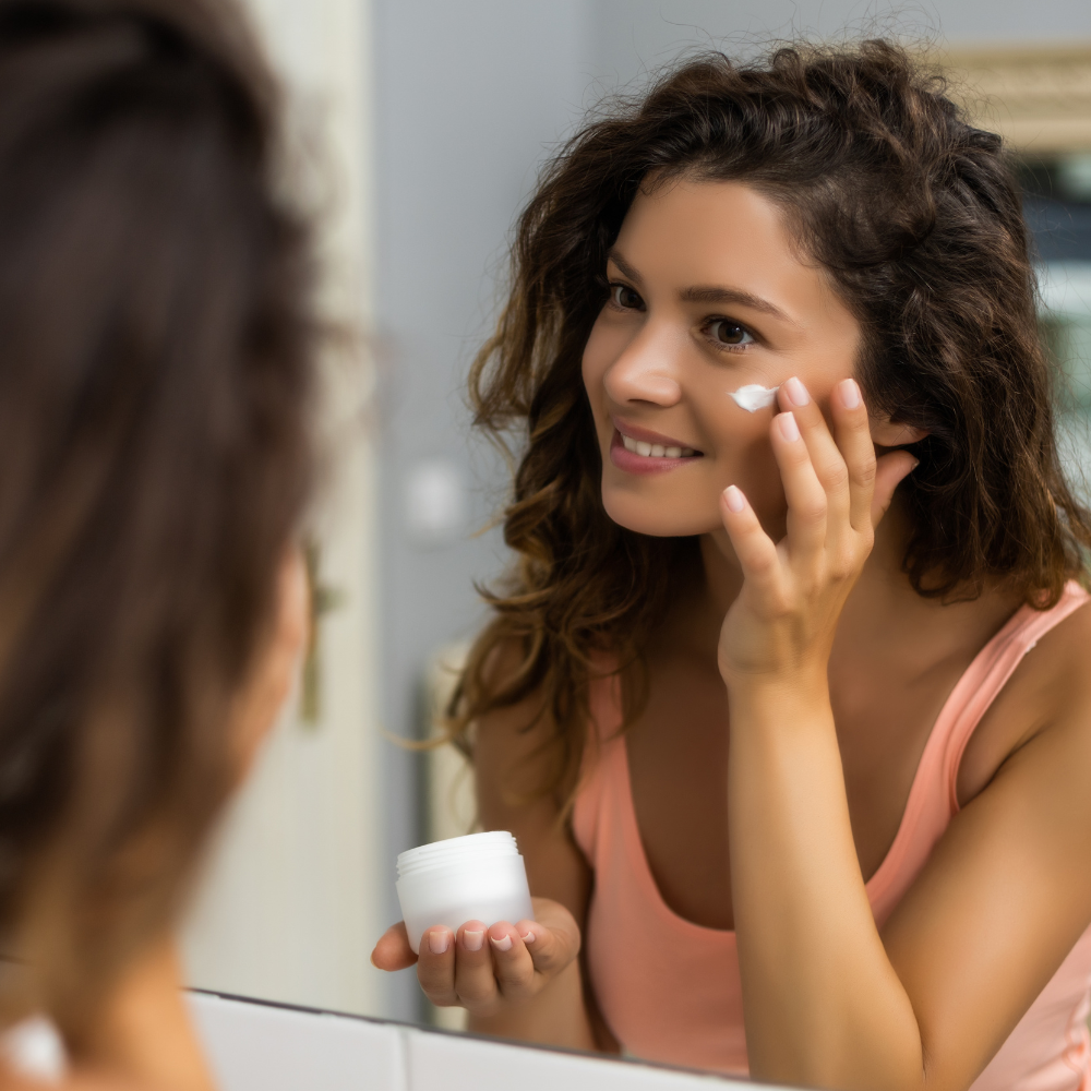 a dark-haired woman applies cream to her face in front of a mirror