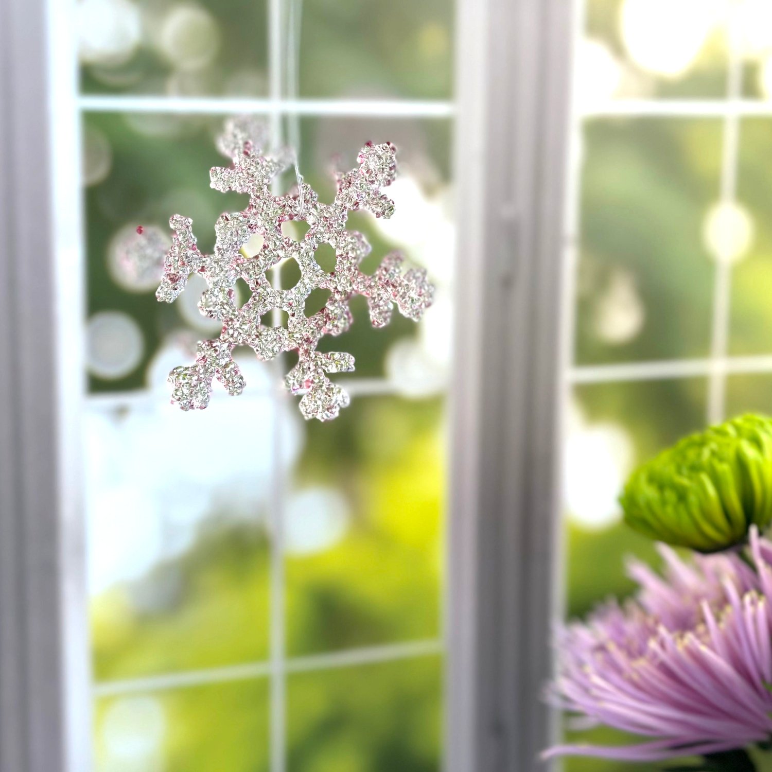 pink glass snowflake hanging in sunny window with purple and green flowers