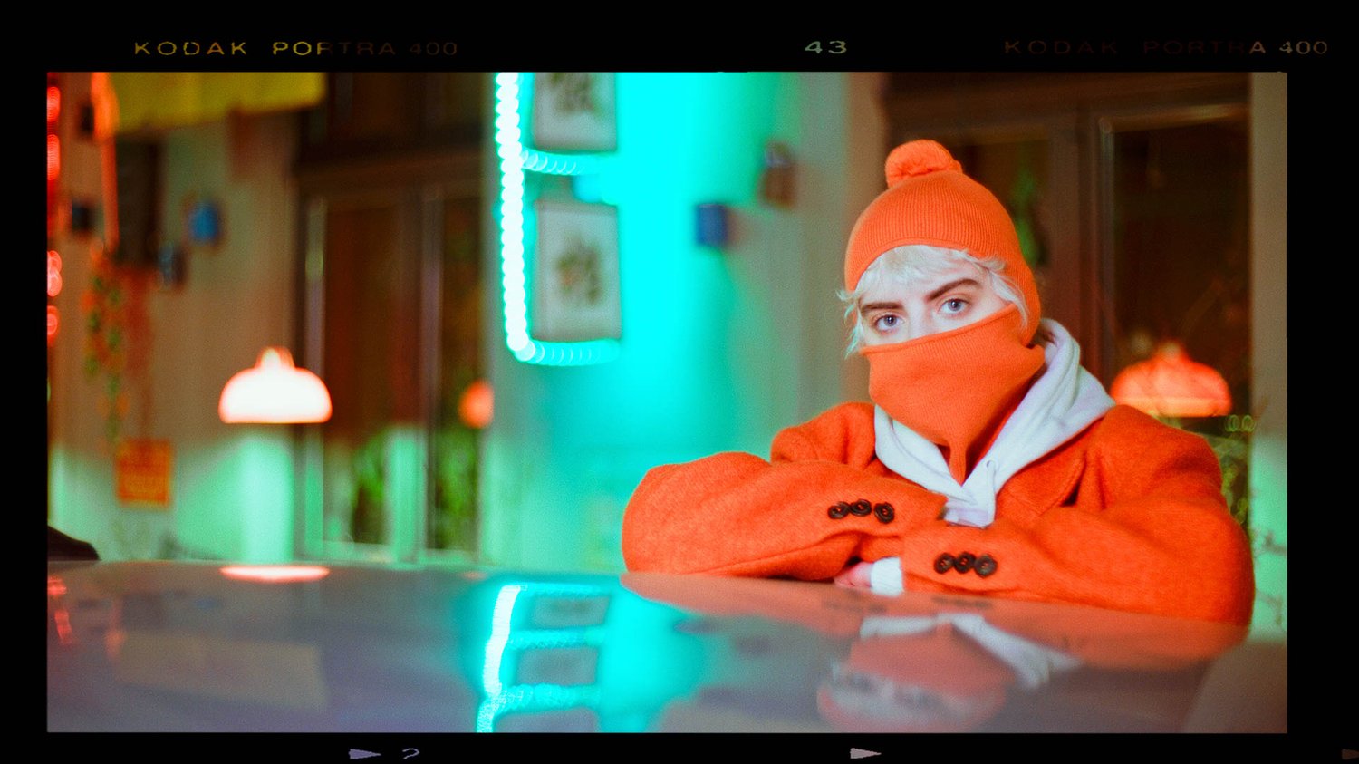 A woman model wears an orange jacket, standing in front of neon light.