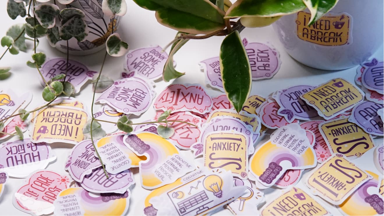 A collection of various stickers scattered around a plant. The stickers include phrases like "Drink Some Water," "I Need A Break," "Anxiety Is," and "ADHD As F*ck," along with a "Rainbow of Disabilities" design.