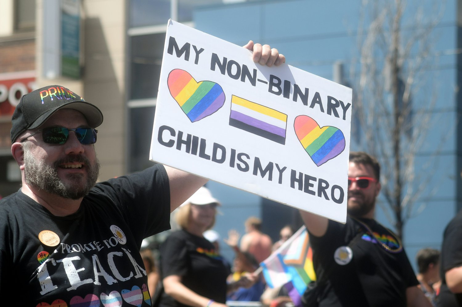 Sign at a PRIDE event with two hearts in rainbow and the trans flag in the middle that reads 'my non-binary child is my hero'