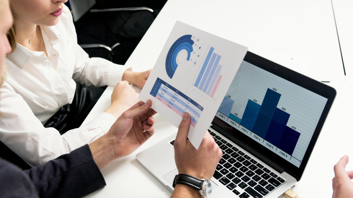 photo of a woman in a white dress shirt and man in black suit jacket looking at data and charts on a print out and a laptop.