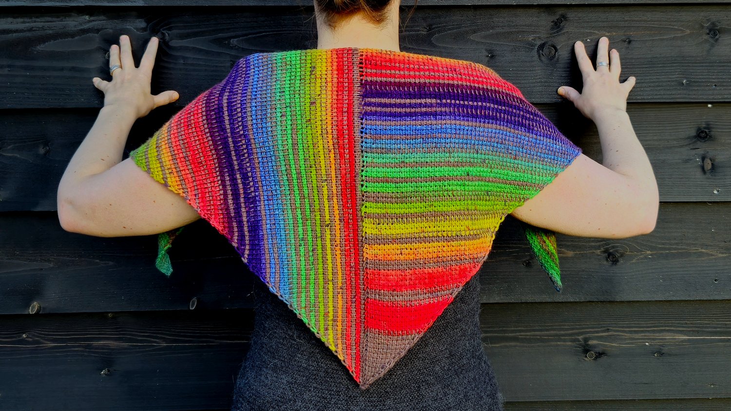 A Tunisian crocheted shawl in rainbow stripes that are vertical on the left side and horizontal on the right side. The shawl is a symmetrical triangle, worn on the back draped over shoulders. The rainbow colored stripes are broken up by a brown stripe bet
