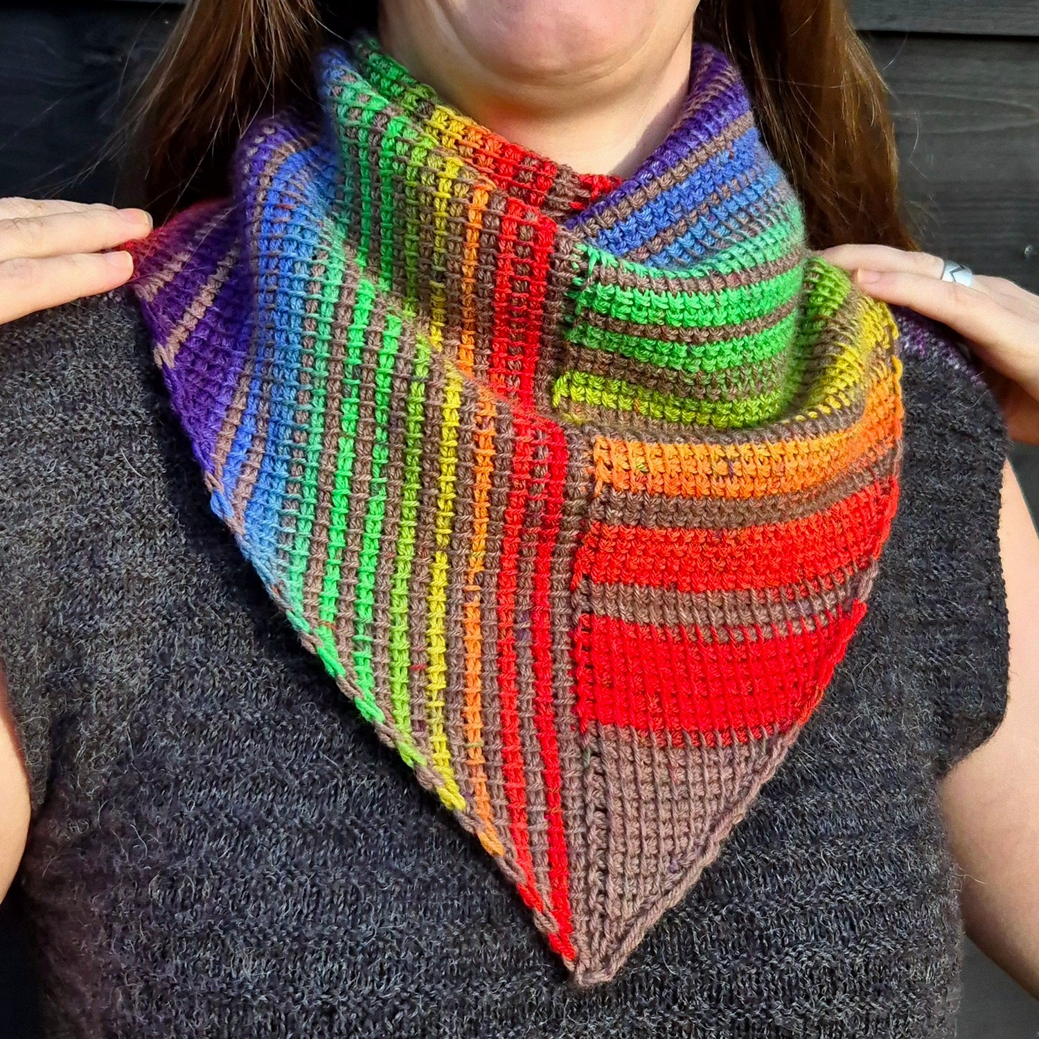 A Tunisian crocheted shawl in rainbow stripes that are vertical on the left side and horizontal on the right side. The shawl is a symmetrical triangle, worn wrapped around the neck with the point in front. The rainbow colored stripes are broken up by a br