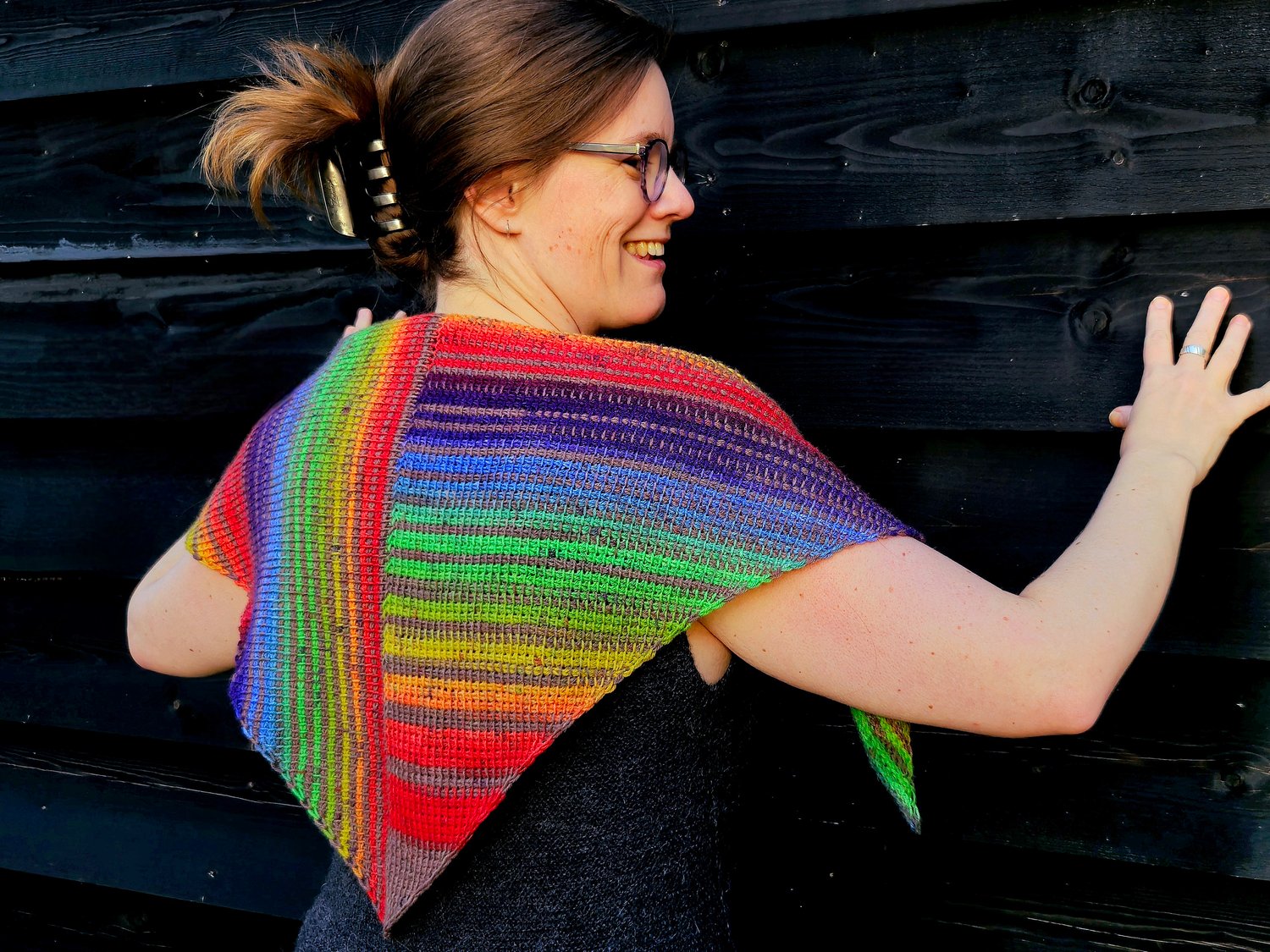 A Tunisian crocheted shawl in rainbow stripes that are vertical on the left side and horizontal on the right side. The shawl is a symmetrical triangle, worn on the back draped over shoulders. The rainbow colored stripes are broken up by a brown stripe bet