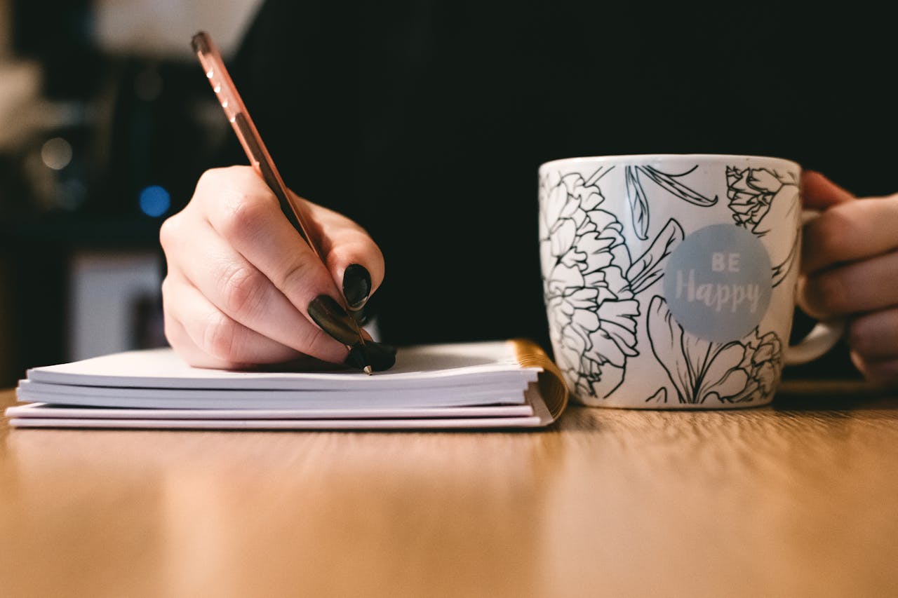 Image of a woman's hands. She is writing on a notebook with one hand and holding a coffee cup in the other. The cup says 