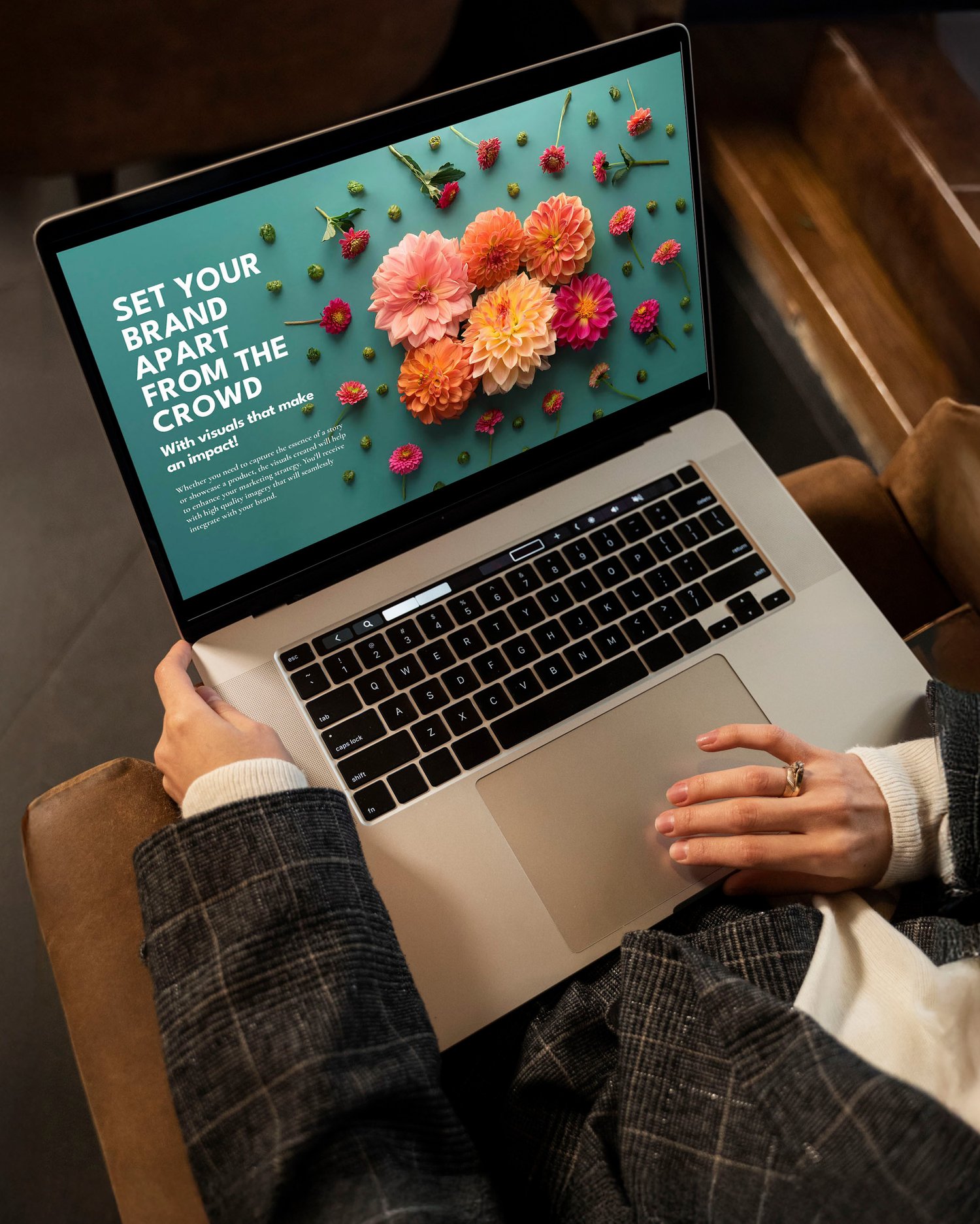 Person seated with a laptop on their lap, displaying a floral design with pink and orange flowers on a teal background. Text on the screen reads: SET YOUR BRAND APART FROM THE CROWD. The person wears a gray plaid blazer.