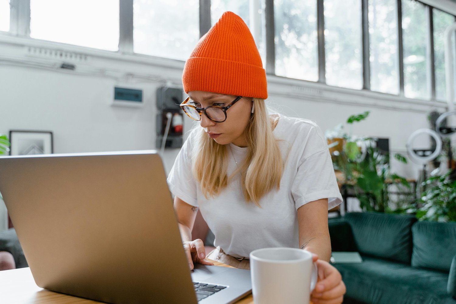 A person wearing an orange beanie and glasses sits at a table, focused on a laptop. They hold a white mug. The room has large windows and plants, creating a bright and relaxed atmosphere.