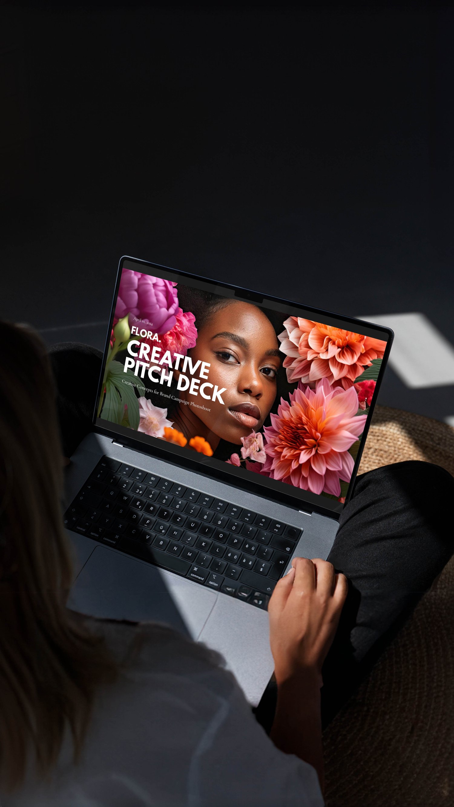 Person sitting with a laptop displaying a creative pitch deck cover featuring a womans face surrounded by colorful flowers. Soft lighting casts shadows on the floor.