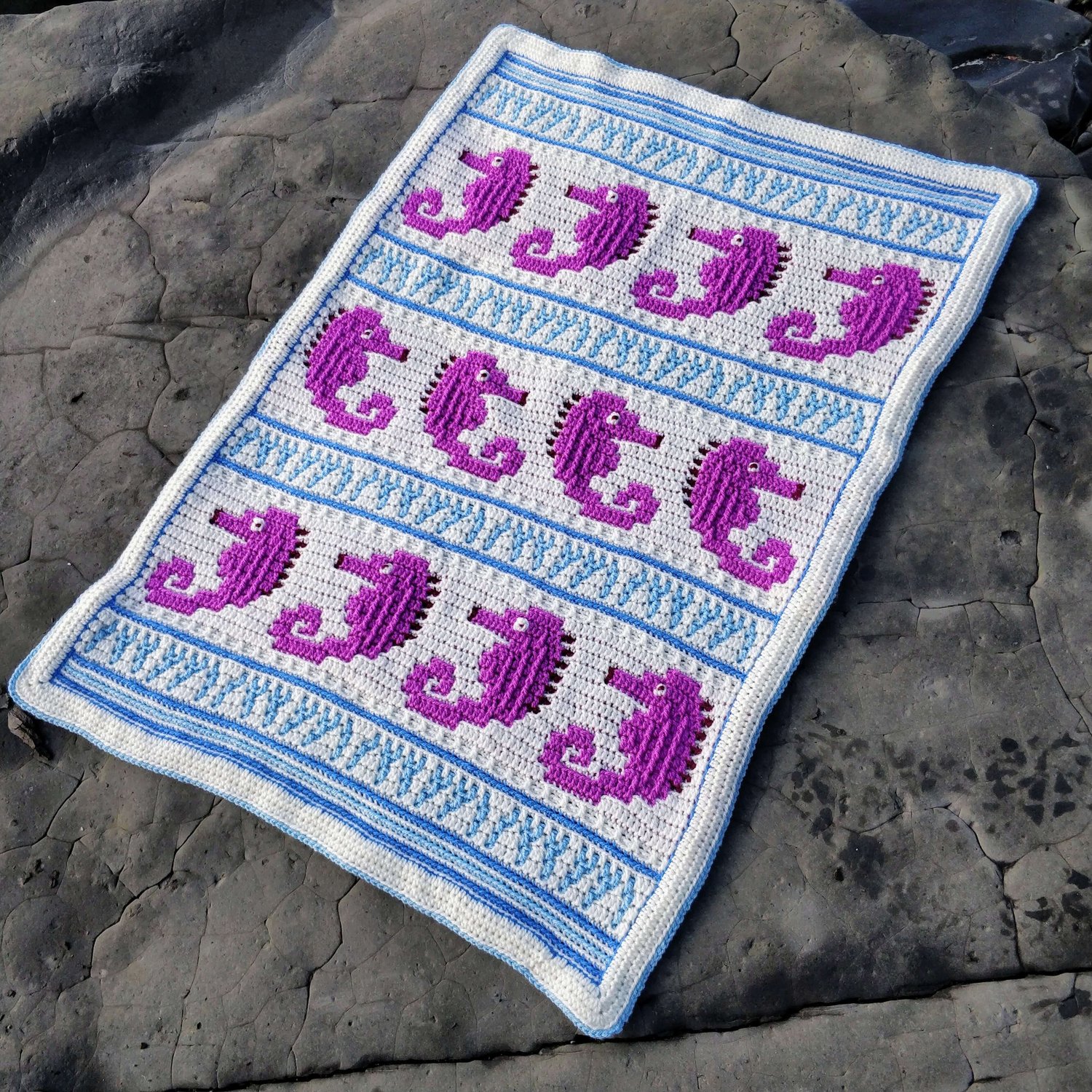 Crocheted blanket with pink seahorses in stripes separated by waves laid on a rock