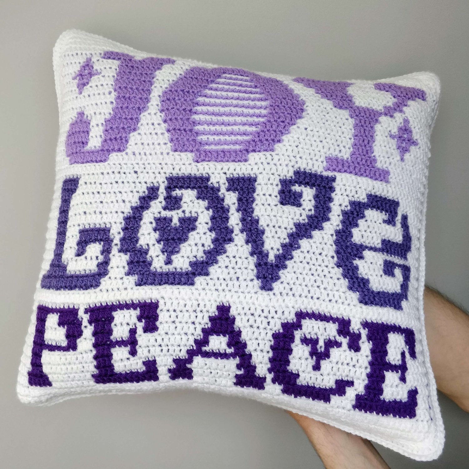 Crochet cushion with Joy Love Peace across it in purples