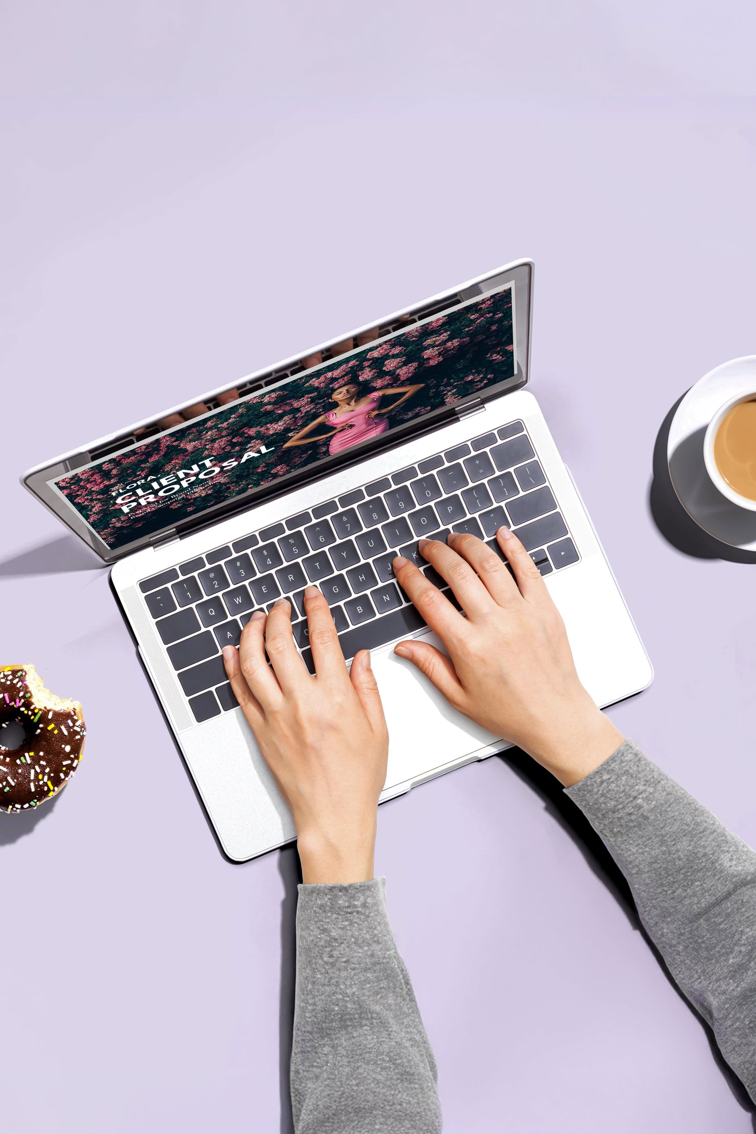 A person typing on a laptop displaying colorful graphics. A chocolate donut and a cup of coffee are on the table beside the laptop. The background is a soft purple.