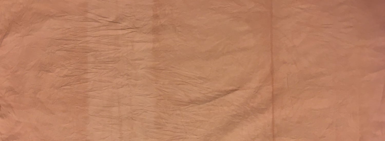 A stainless steel pot on a stove, filled with fabric immersed in a rich, maroon-colored natural dye made from avocado pits and skins. The fabric is evenly soaked, with folds visible above the liquid.