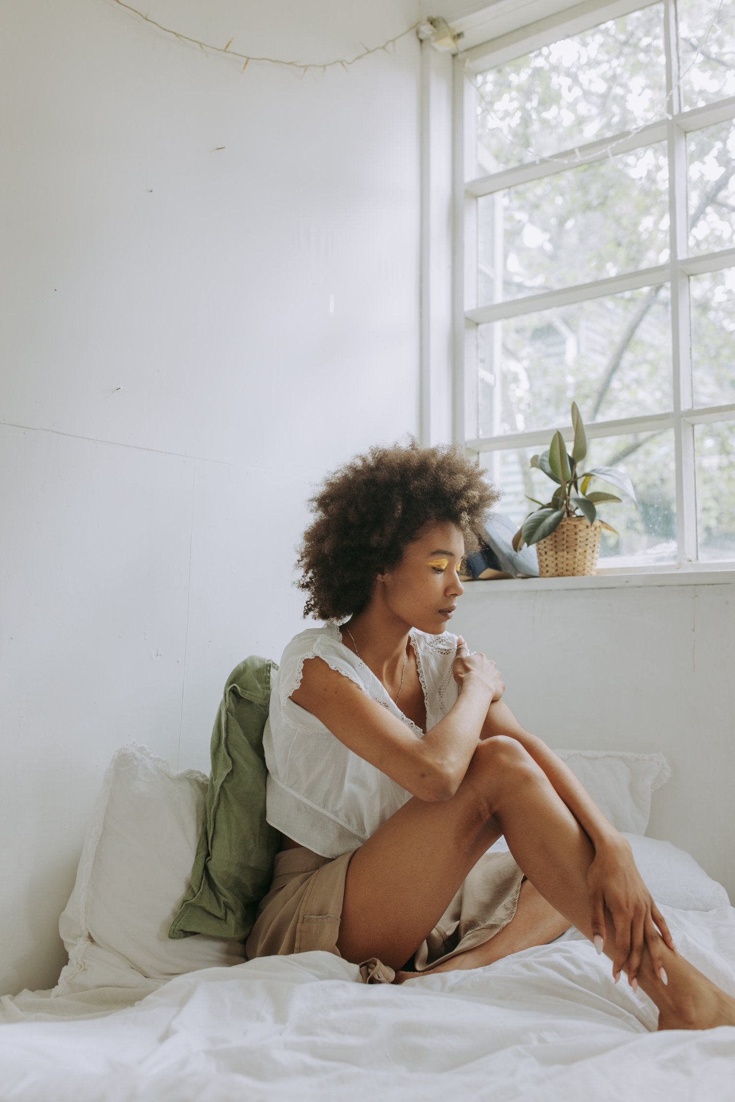Contemplative Young Black Woman Sitting In Her Bed With Sunlight Streaming Through the Window